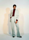 A model stands against a light gray studio background wearing a pale mint green double-breasted suit with wide-leg trousers, paired with a black shirt and black shoes.