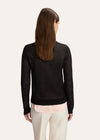 A model wearing a black long-sleeve knit sweater with a light pink collar and 'Fairytale of New York' embroidered in gold on the chest, styled with a light pink collared shirt and cream trousers against a light gray background.