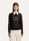A model wearing a black long-sleeve knit sweater with a light pink collar and 'Fairytale of New York' embroidered in gold on the chest, styled with a light pink collared shirt and cream trousers against a light gray background.