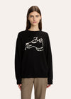 A woman modeling a black crewneck sweater with a white hand-drawn dog graphic on the chest, paired with beige plaid trousers, against a solid off-white background.