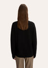 A woman modeling a black crewneck sweater with a white hand-drawn dog graphic on the chest, paired with beige plaid trousers, against a solid off-white background.