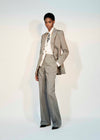 A model stands against a light studio background wearing a tailored light gray pinstripe suit with a white shirt, patterned tie, and black pointed shoes.