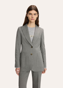 A model wearing a tailored pinstripe blazer over a white collared shirt and dark vest, paired with a matching pleated mini skirt, photographed against a plain light background.